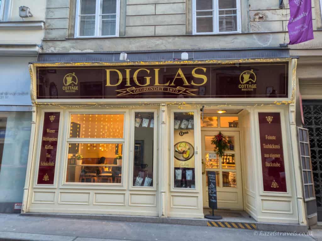 Exterior of Diglas café in Vienna, with a burgundy and gold sign above the entrance, warm lights in the window, and a cream-coloured façade along a narrow street.