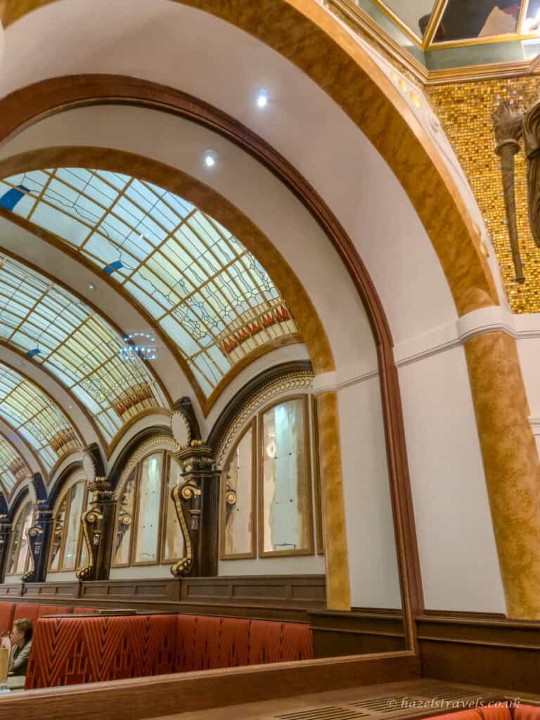 Interior of a grand Vienna café with arched ceilings, decorative columns, patterned glass panels, and red upholstered seating beneath warm lighting.