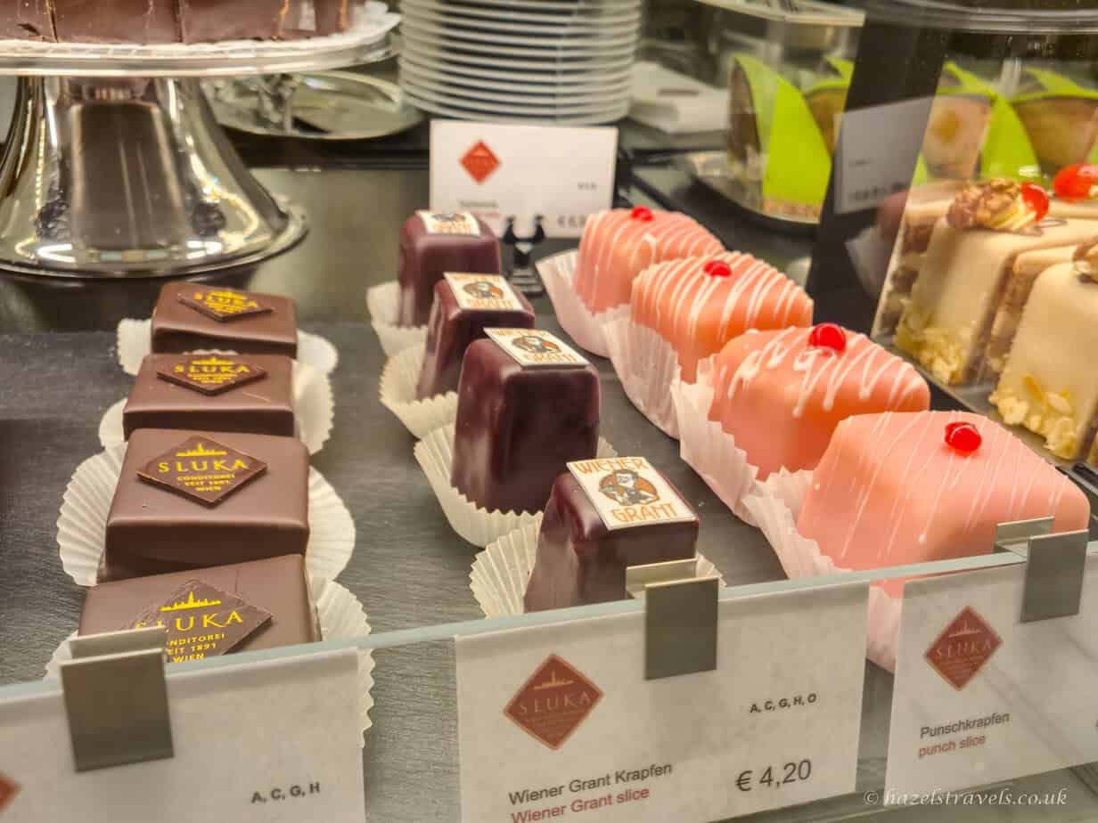 Selection of neatly arranged cakes and slices displayed behind a glass counter, including pink marzipan squares and chocolate-topped pastries with price labels below.