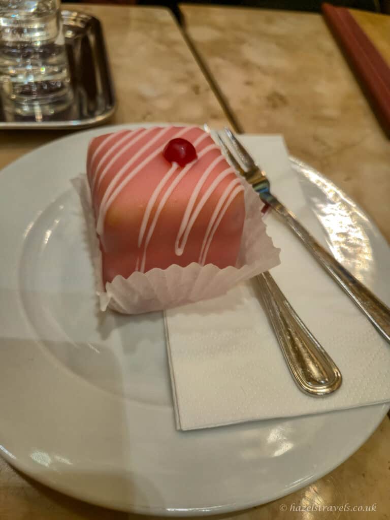 Punschkrapfen, a pink Viennese sponge cake coated in marzipan with white icing stripes and a cherry on top, served on a white plate with a fork and napkin.