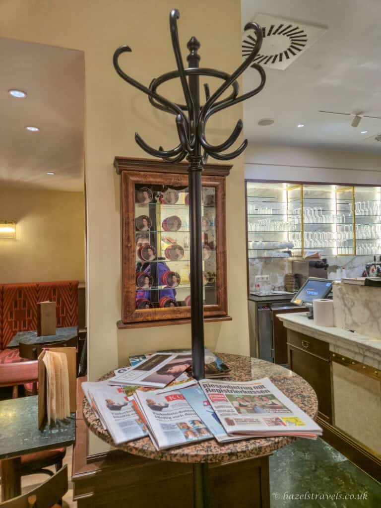 Traditional newspaper stand with wooden coat hooks and daily papers displayed on a round marble table inside a Viennese café, with seating and a counter in the background.