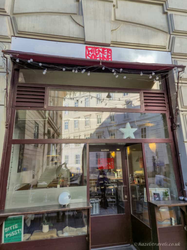 Entrance to Wiener Söhne café in Vienna, with a glass front door, large windows reflecting nearby buildings, and a red sign above the doorway.