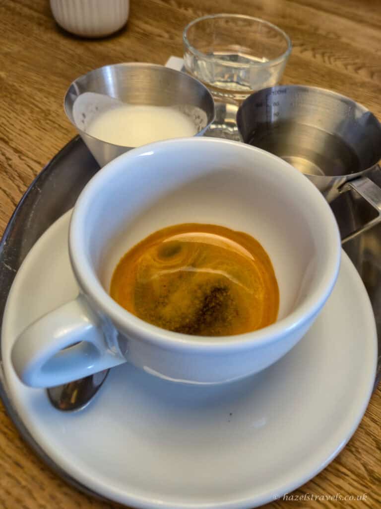 Cup of espresso served on a saucer with separate metal pitchers of milk and hot water, and a glass of water on a wooden table.