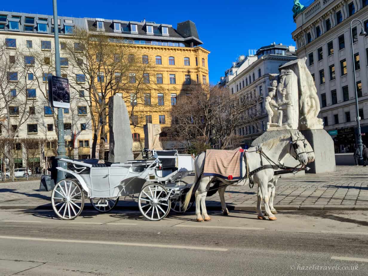 White horse-drawn carriages waiting in a Vienna city square, with historic cream and yellow buildings and a large stone monument in the background under a clear blue sky.