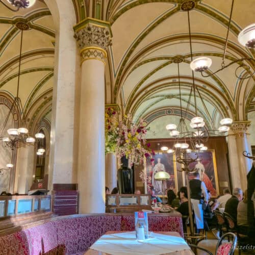 Interior of Café Central in Vienna with cream and green vaulted ceilings, marble columns, ornate chandeliers and diners seated at red-upholstered tables beneath large portraits.