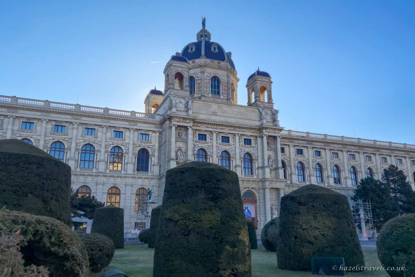 Kunsthistorisches Museum in Vienna with its domed roof and ornate cream stone façade, framed by neatly trimmed green hedges under a pale blue sky.