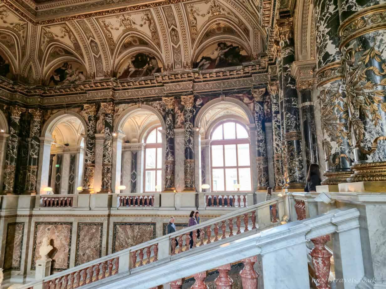Ornate interior of the Kunsthistorisches Museum in Vienna, with dark green and pink marble columns, gilded ceilings, arched windows and a sweeping stone staircase.