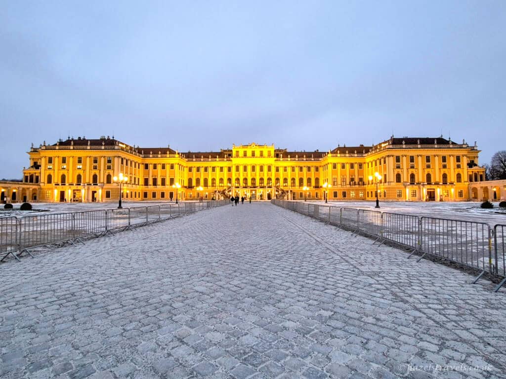 Schönbrunn Palace in Vienna illuminated at dusk, with golden lights reflecting on the grand Baroque façade and a snow-dusted cobbled courtyard in the foreground.