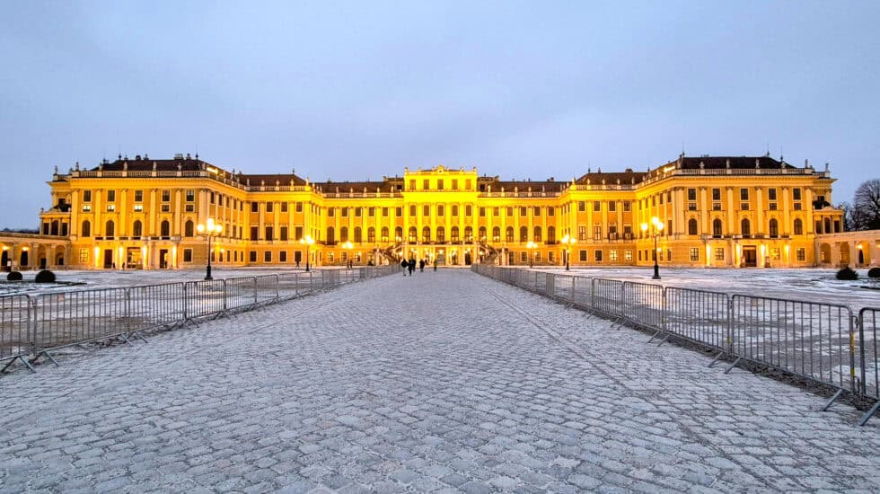 Schönbrunn Palace in Vienna illuminated at dusk, with golden lights reflecting on the grand Baroque façade and a snow-dusted cobbled courtyard in the foreground.
