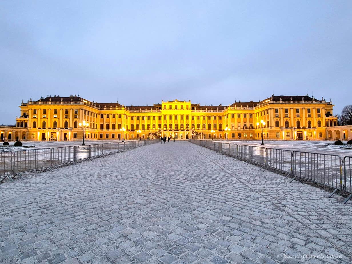 Schönbrunn Palace in Vienna illuminated at dusk, with golden lights reflecting on the grand Baroque façade and a snow-dusted cobbled courtyard in the foreground.