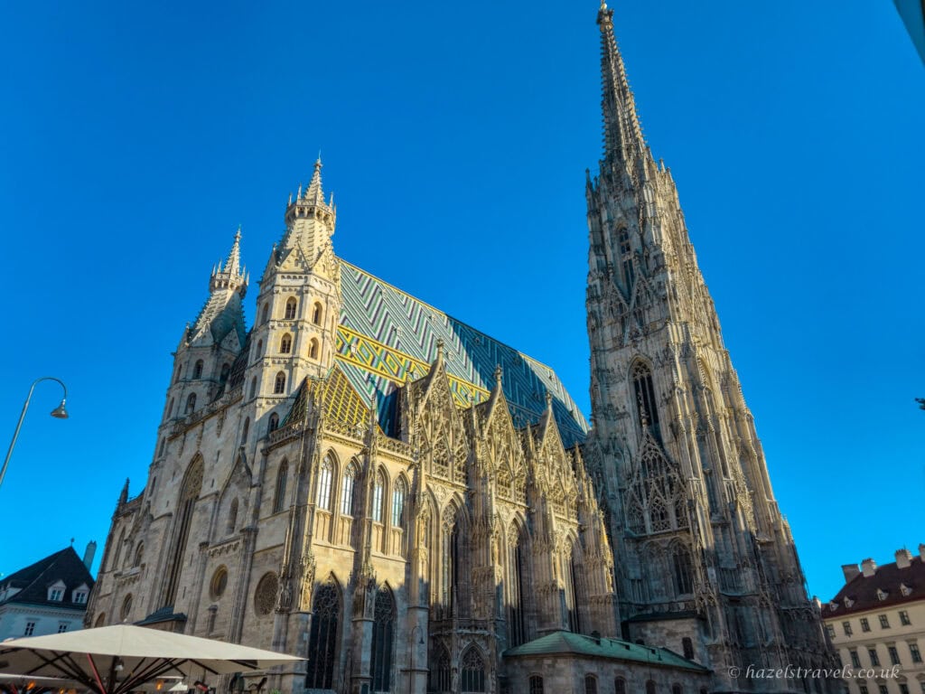 St Stephen’s Cathedral in Vienna with its tall Gothic spire and patterned green, yellow and blue tiled roof under a clear blue sky.