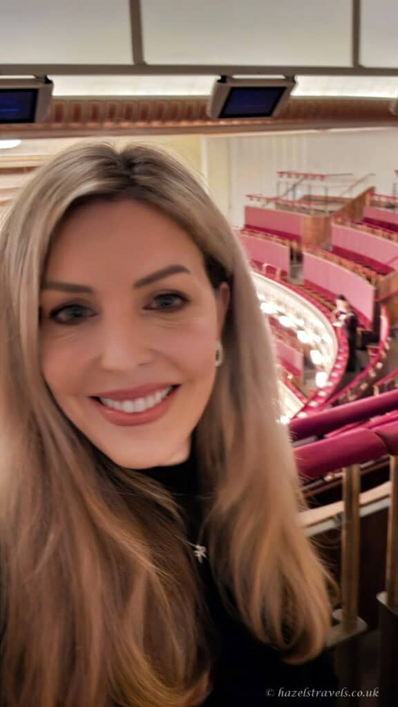 Smiling woman taking a selfie inside the Vienna State Opera, with curved tiers of pink and red balcony seating visible in the background.