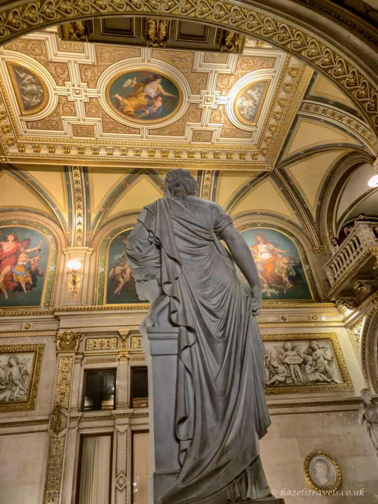Grand interior of the Vienna State Opera with ornate arches, gilded detailing, ceiling frescoes, and a marble statue standing beneath the decorated vaulted ceiling.