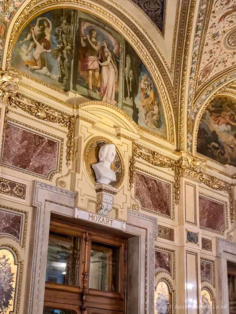 Ornate interior of the Vienna State Opera with gold detailing, rose and cream marble panels, painted ceiling frescoes, and a white marble bust of Mozart above a dark wooden doorway.