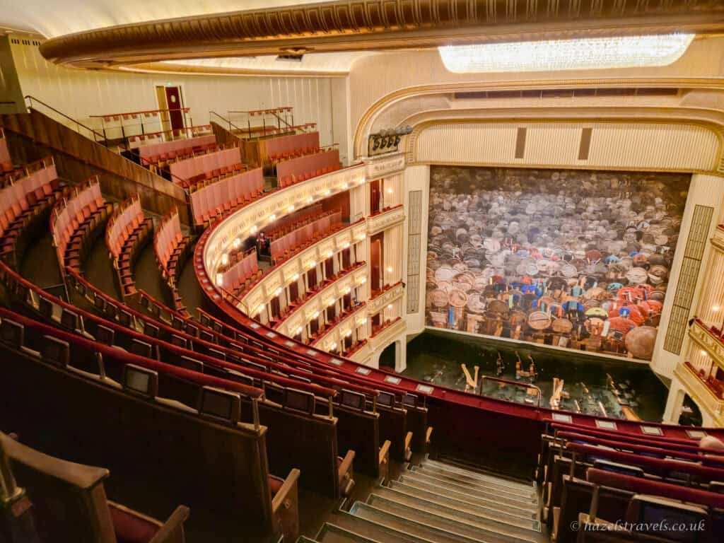 Auditorium of the Vienna State Opera with curved tiers of red and pink seats, cream and gold balconies, and a large stage curtain featuring an image of a crowd scene.
