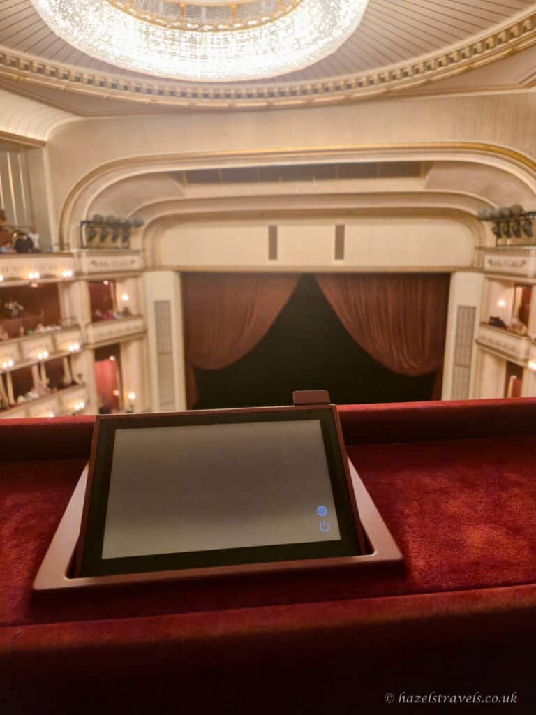 View from a red velvet balcony inside the Vienna State Opera, showing cream and gold tiers of boxes, a crystal ceiling light, closed red stage curtains, and a small digital screen in the foreground.