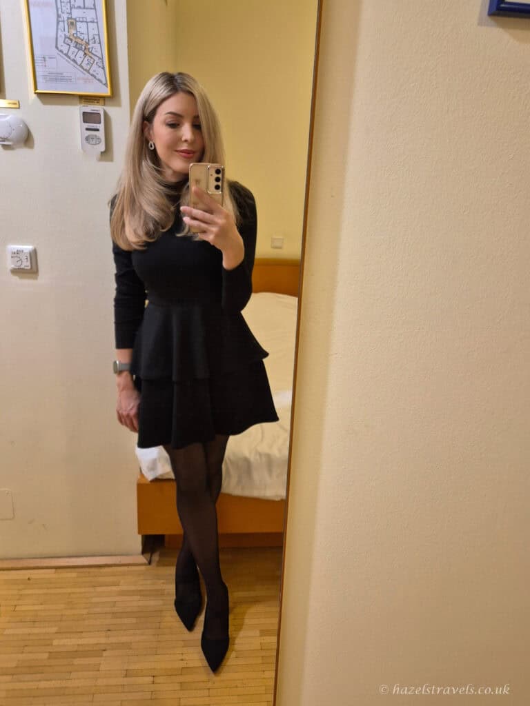 Woman taking a mirror selfie in a hotel room, wearing a fitted black long-sleeve dress with a flared skirt, black tights, and pointed heels.
