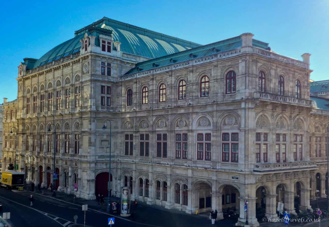 Exterior of the Vienna State Opera building in pale stone, with arched windows, ornate detailing and a green copper roof, photographed in soft evening light from across the street.