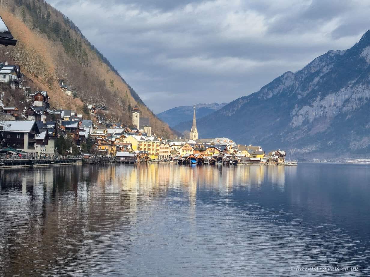 Hallstatt village reflected in the calm waters of Lake Hallstatt, with colourful lakeside houses and a tall church spire beneath dramatic Alpine mountains.