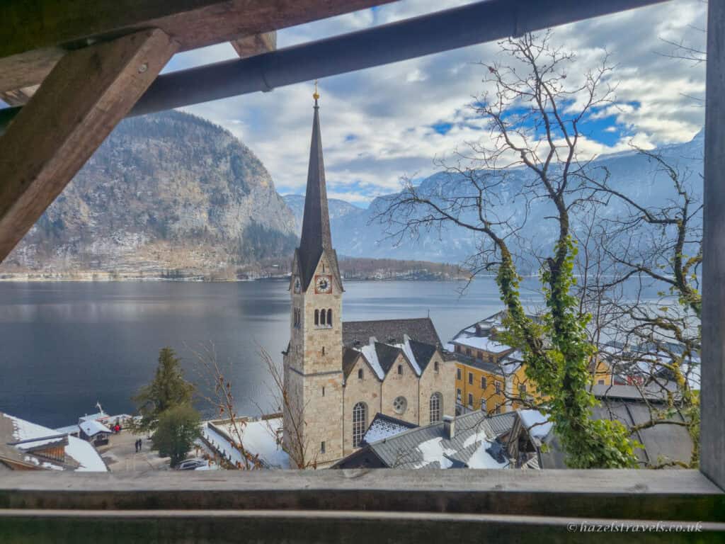 View of Hallstatt’s Evangelical Church spire beside Lake Hallstatt, framed by a wooden balcony with snowy rooftops and mountains in the background.