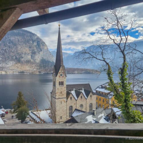 View of Hallstatt’s Evangelical Church spire beside Lake Hallstatt, framed by a wooden balcony with snowy rooftops and mountains in the background.