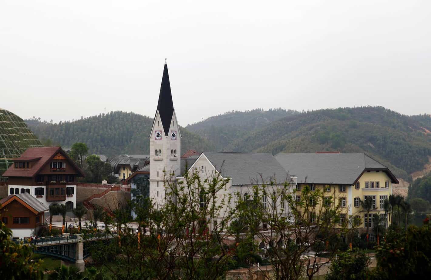 Replica of the Austrian village of Hallstatt in Huizhou, China, with a white church tower and Alpine-style buildings surrounded by low green hills.