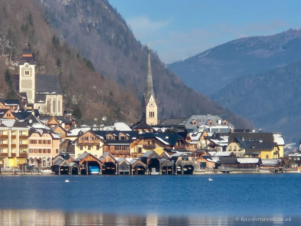 Colourful lakeside houses in Hallstatt reflected in Lake Hallstatt, with church spires rising above the village and forested mountains in the background.