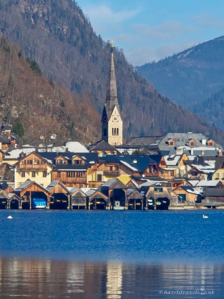 Colourful lakeside houses in Hallstatt reflected in Lake Hallstatt, with church spires rising above the village and forested mountains in the background.
