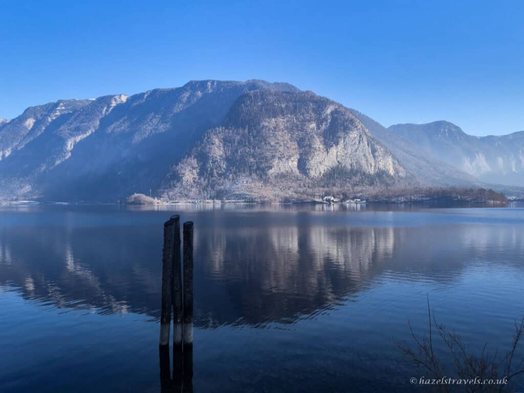 Calm water of Lake Hallstatt reflecting a large mountain under a clear blue sky, with two wooden posts standing in the lake in the foreground.