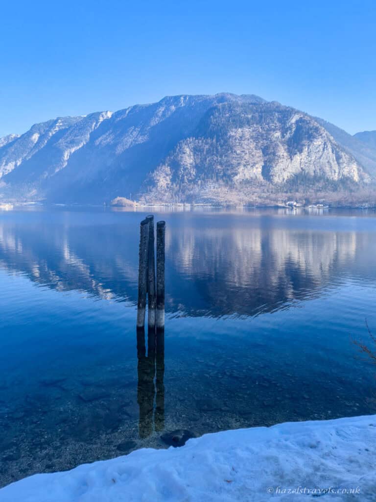 Calm water of Lake Hallstatt reflecting a large mountain under a clear blue sky, with two wooden posts standing in the lake in the foreground.