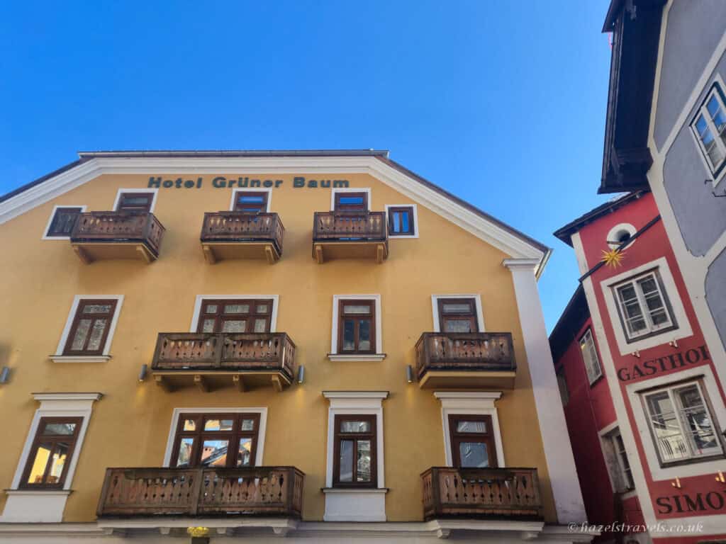 Yellow building of Hotel Grüner Baum in Hallstatt with wooden balconies and windows, standing beside a red alpine building under a clear blue sky.