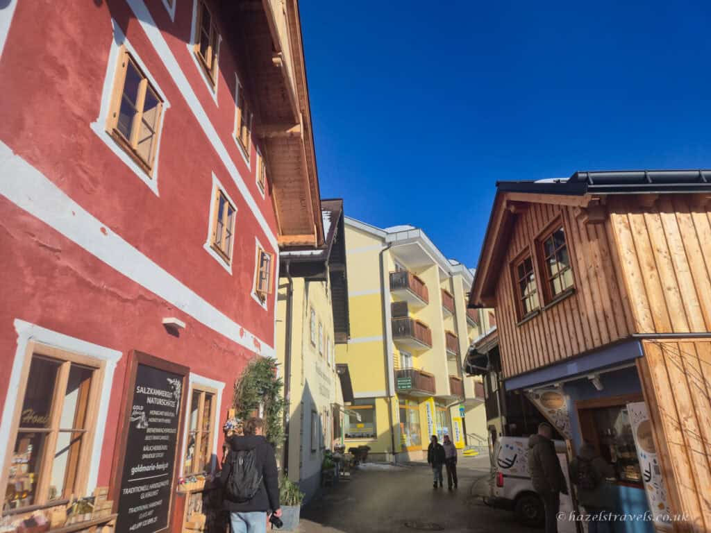 Narrow street in Hallstatt lined with colourful alpine buildings, including a red shopfront and wooden house, with people walking under a clear blue sky.