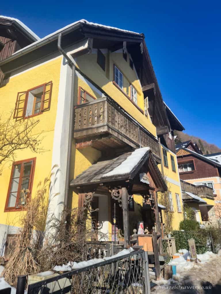 Yellow alpine house in Hallstatt with a wooden balcony and carved wooden porch above a snowy walkway under a clear blue sky.
