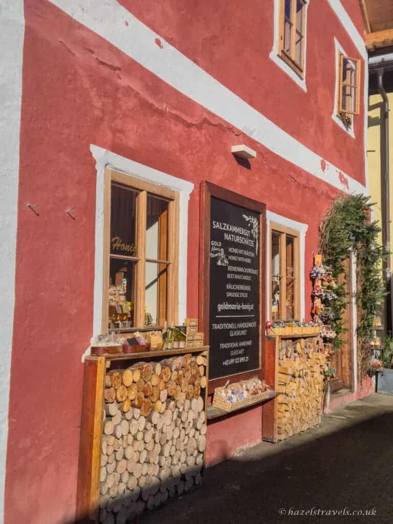 Red shopfront in Hallstatt with wooden windows displaying souvenirs and a sign advertising local Salzkammergut products, with stacked firewood beneath the windows.