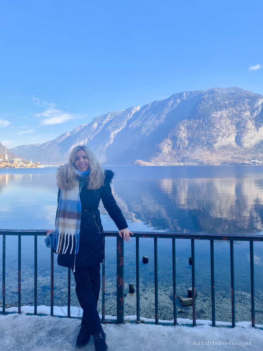 Woman standing by a lakeside railing at Lake Hallstatt, with calm water reflecting snow-covered Alpine mountains under a bright blue winter sky.