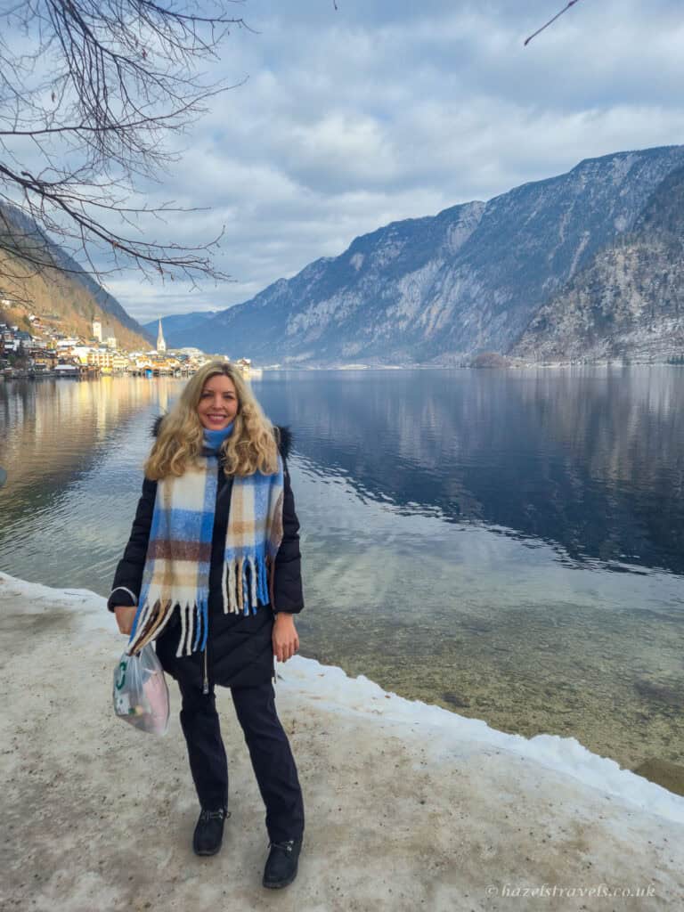 Woman standing beside Lake Hallstatt on a snowy winter day, with calm water reflecting the Alpine mountains and the village visible in the distance.