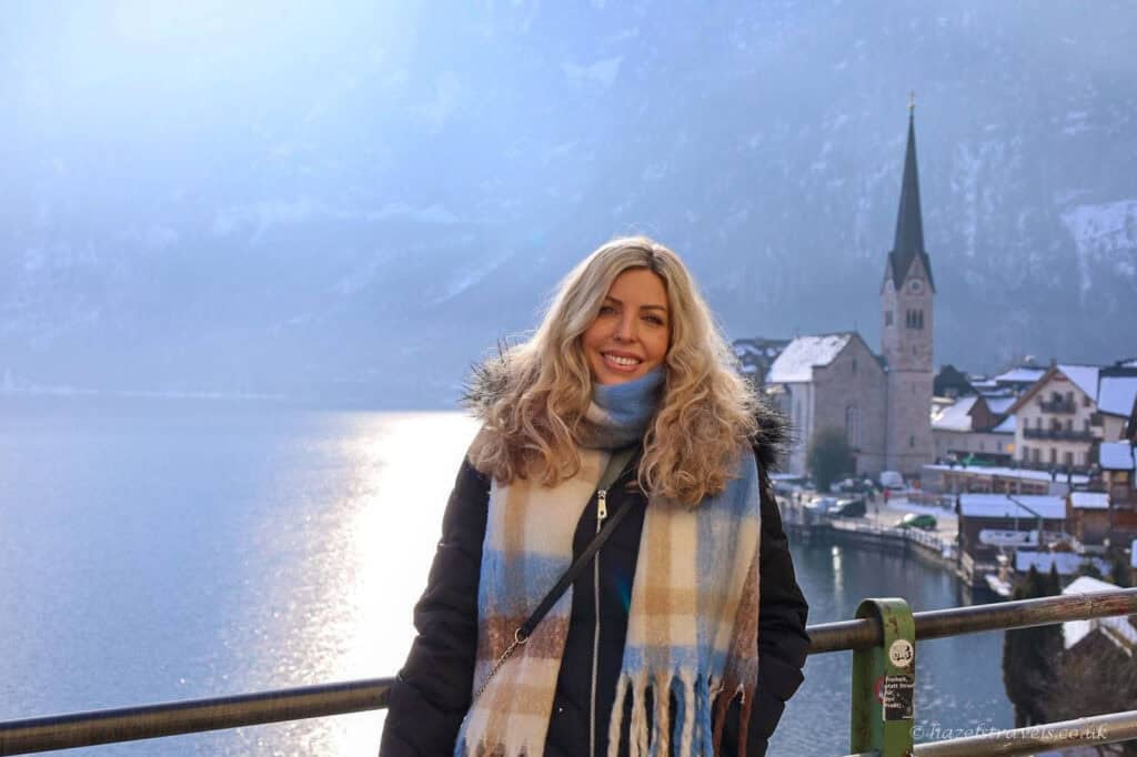 Woman standing beside Lake Hallstatt on a snowy winter day, with calm water reflecting the Alpine mountains and the village visible in the distance.