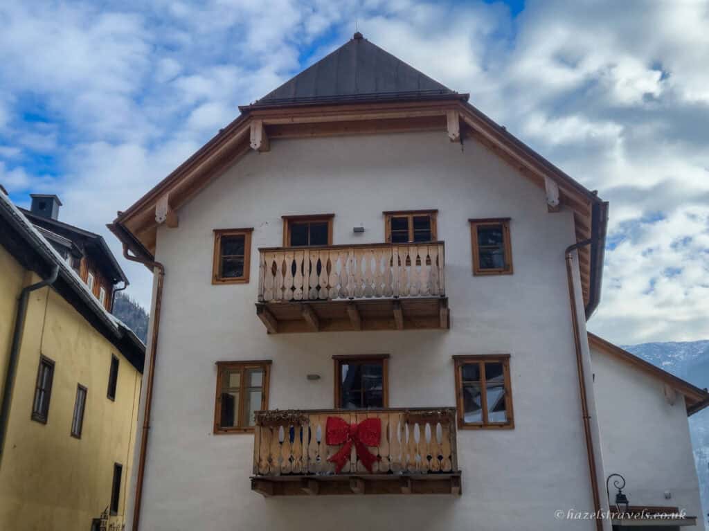 White alpine house in Hallstatt with wooden balconies and a red decorative bow on the lower balcony, under a cloudy blue sky.