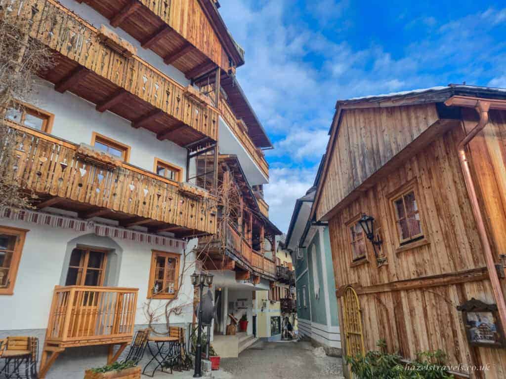Narrow street in Hallstatt lined with alpine houses featuring wooden balconies and timber facades under a partly cloudy blue sky.