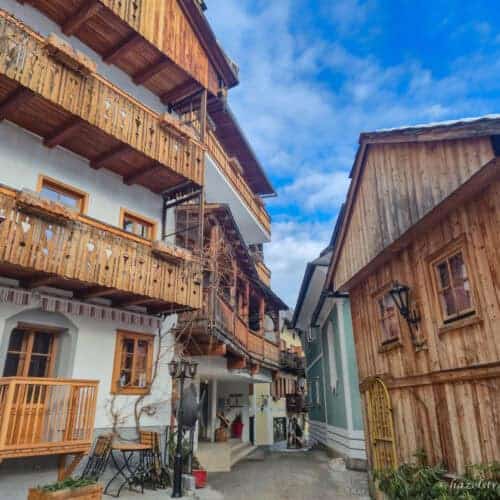 Narrow street in Hallstatt lined with alpine houses featuring wooden balconies and timber facades under a partly cloudy blue sky.
