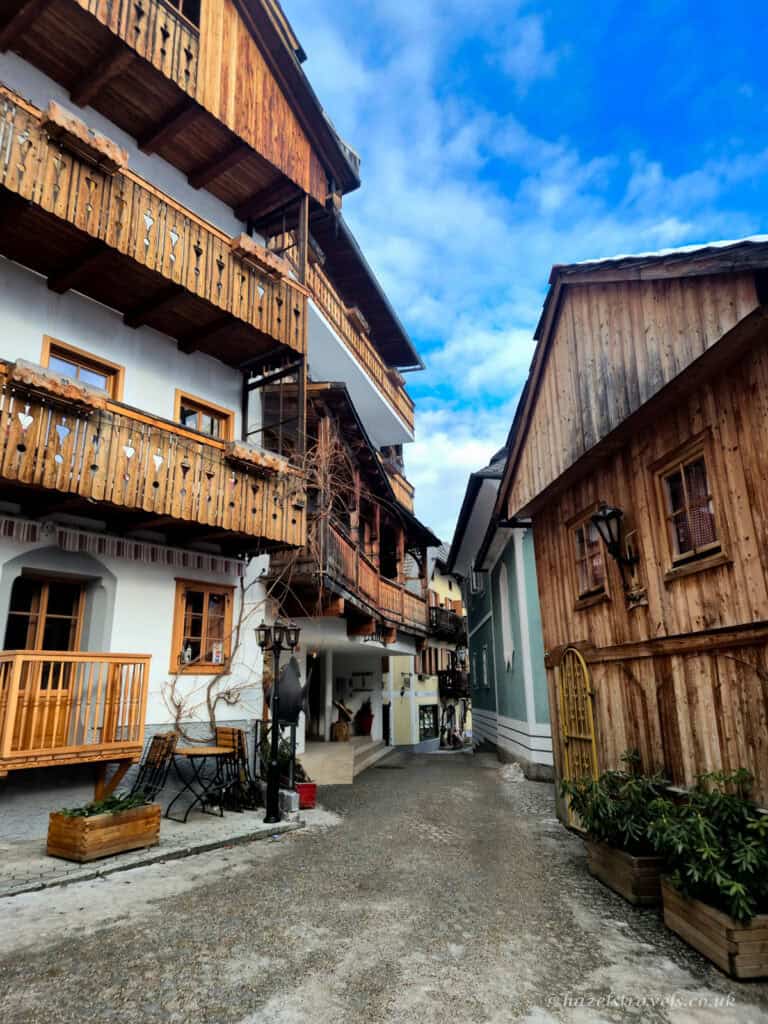 Narrow street in Hallstatt lined with alpine houses featuring wooden balconies and timber facades under a partly cloudy blue sky.