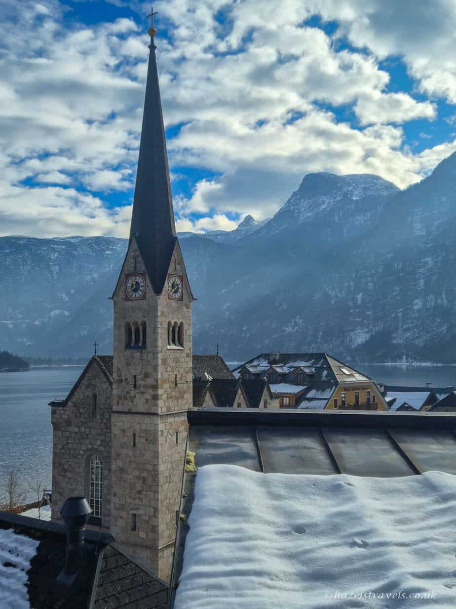 Tall church spire of Hallstatt’s Evangelical Church rising above snowy rooftops beside Lake Hallstatt, with mountains and clouds in the background.
