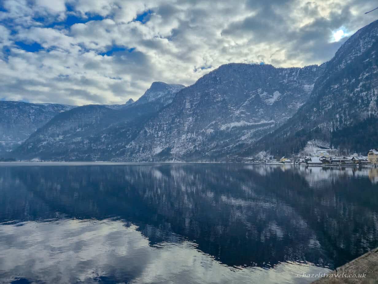 Calm blue water of Lake Hallstatt reflecting cloudy winter skies, surrounded by steep snow-dusted mountains in the Austrian Alps.
