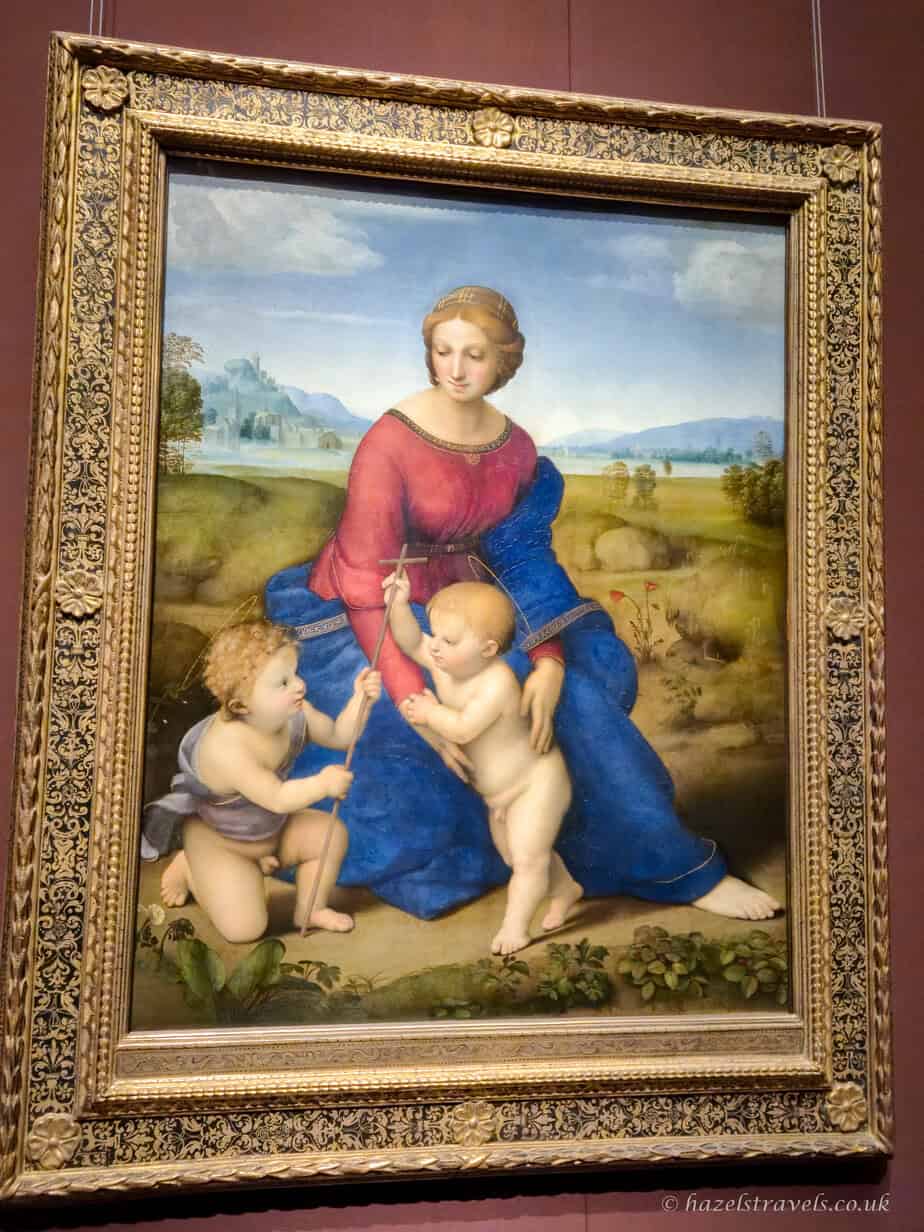 The painting “Madonna of the Meadow” by Raphael depicting Mary with the infant Jesus and John the Baptist in a peaceful countryside setting.