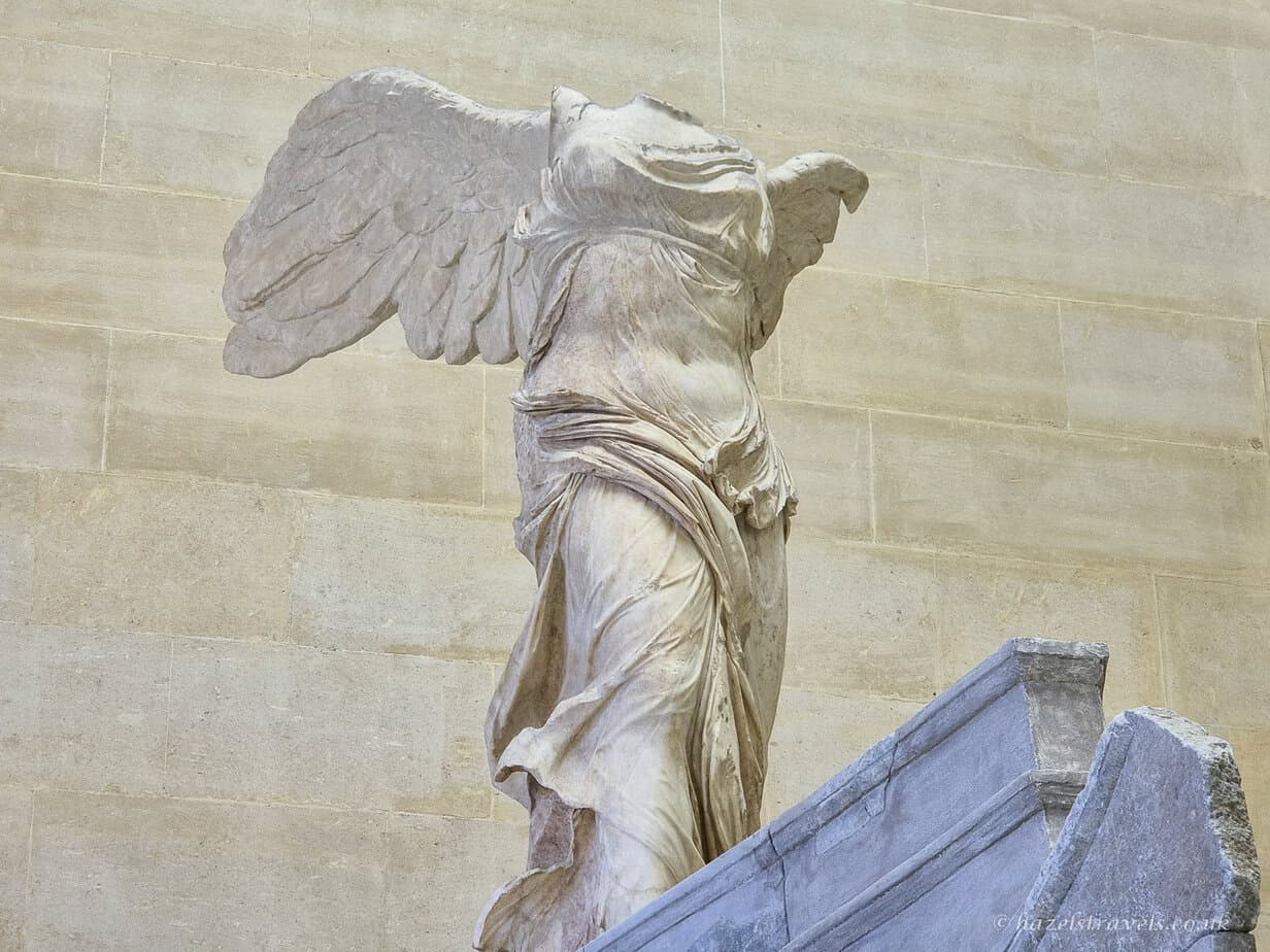 Winged Victory of Samothrace with visitors, Louvre Museum — the iconic ancient sculpture of Nike seen from below looking up