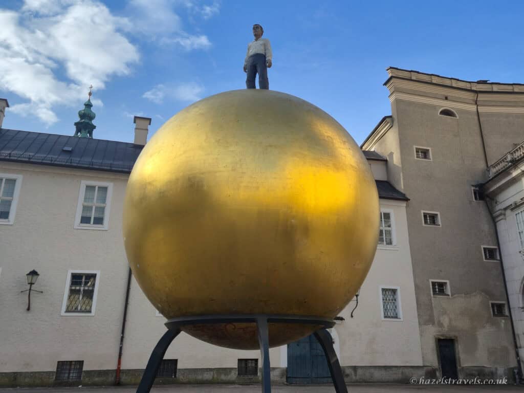 Golden Sphaera sculpture in Kapitelplatz Salzburg with a large gold sphere and a man standing on top, set against pale historic buildings and blue sky