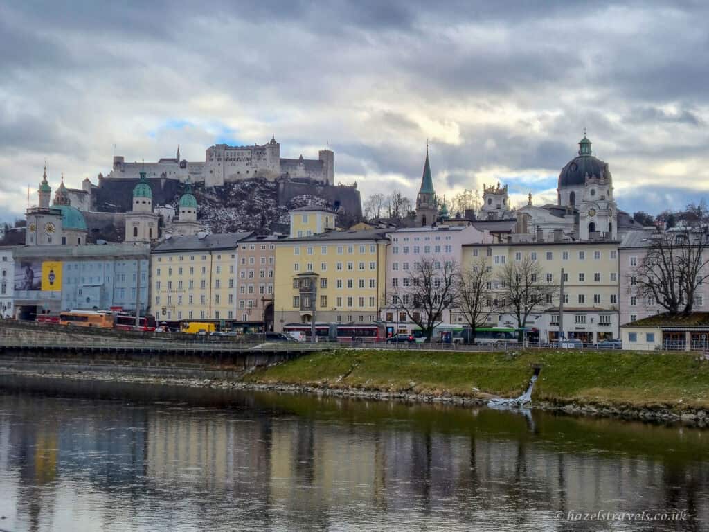 Pastel buildings along the Salzach River in Salzburg with church domes and Hohensalzburg Fortress on the hill above under a cloudy sky
