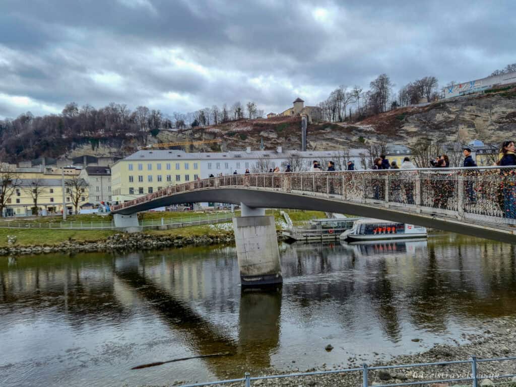 Makartsteg pedestrian bridge in Salzburg covered with colourful love locks spanning the Salzach River, with people crossing and hills in the background under a cloudy sky