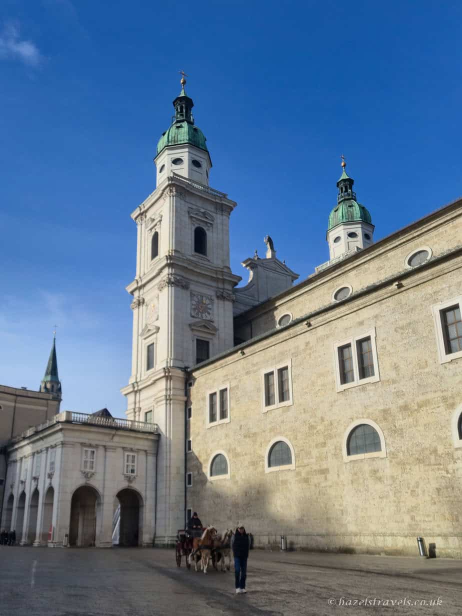 Salzburg Cathedral with pale stone faƧade and twin towers topped with green domes, seen from the square under a clear blue sky with a horse-drawn carriage passing