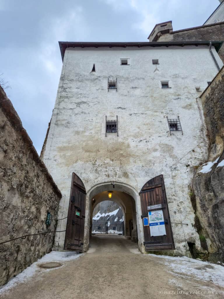 Stone entrance gate to Hohensalzburg Fortress in Salzburg with white fortress walls, wooden doors, and snowy hillside visible through the archway